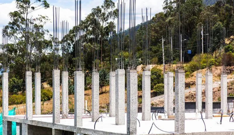 Fila de columnas de concreto recién desencofradas con fierros de espera sobre una losa, con un cerro arbolado al fondo