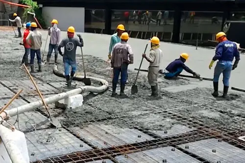 Cuadrilla con cascos amarillos extiende concreto fresco sobre un techo armado mientras una manguera de bombeo descarga la mezcla