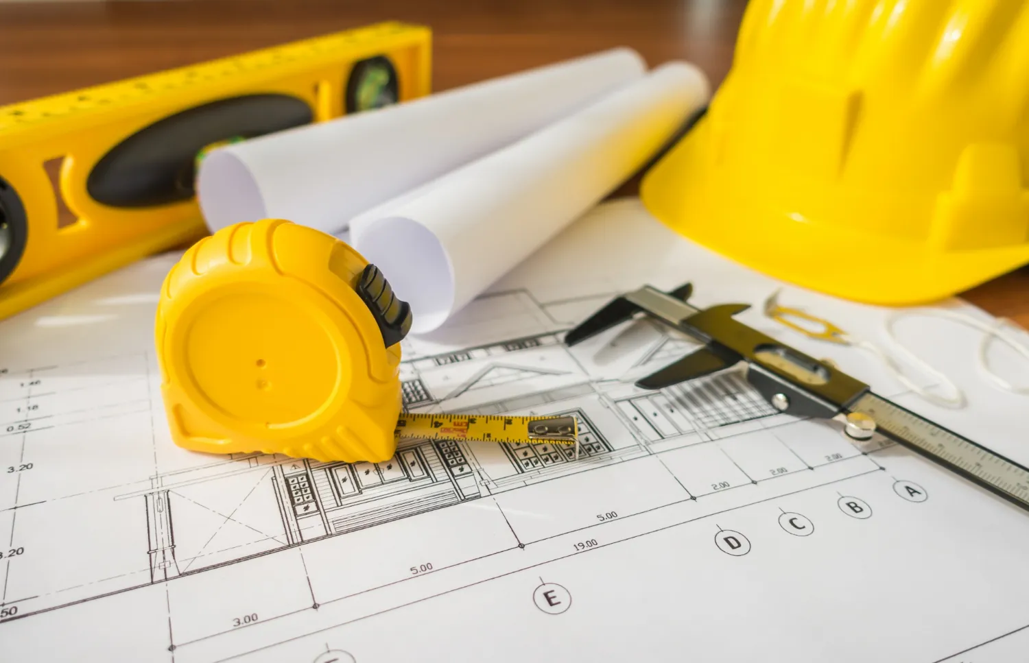 Planos de construcción extendidos sobre una mesa con un casco amarillo, una wincha, un nivel y un calibrador