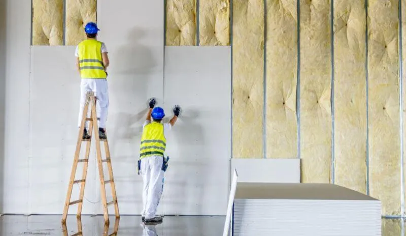 Dos operarios con casco y chaleco colocan planchas de drywall sobre perfiles metálicos, con lana aislante amarilla entre los parantes
